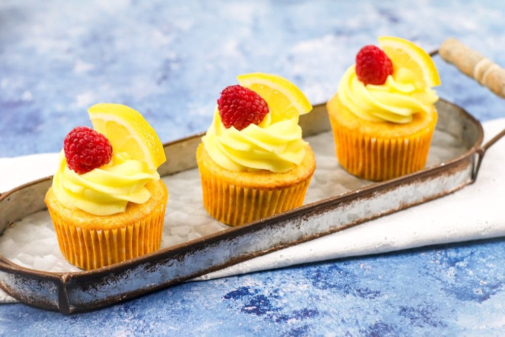 Three lemon cupcakes with yellow frosting and topped with a lemon slice and a raspberry, presented on a metal tray against a blue background.