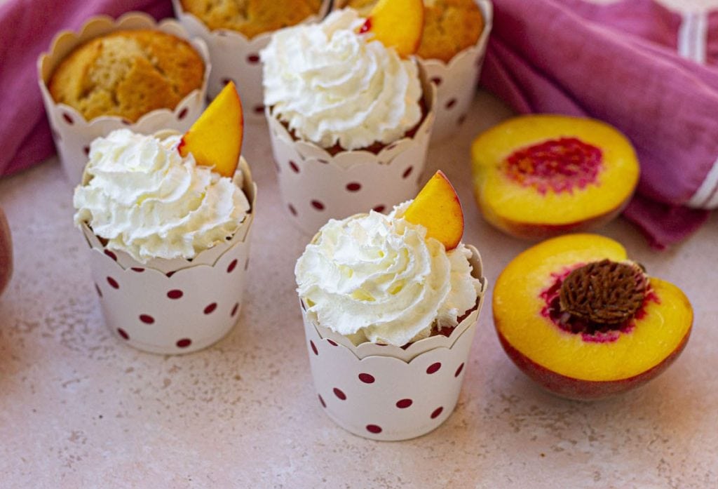 Peach cupcakes topped with whipped cream and a slice of peach, presented in polka-dot cups.