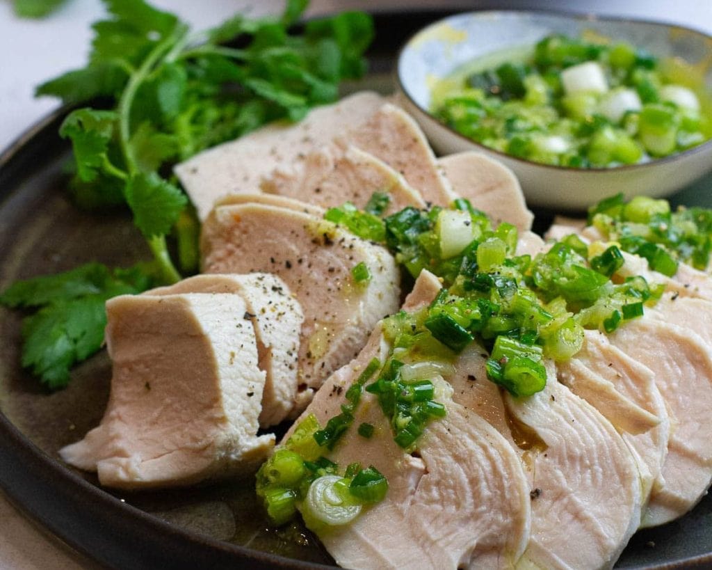 Sliced poached chicken breast topped with scallions and served with a side of dipping sauce and cilantro.