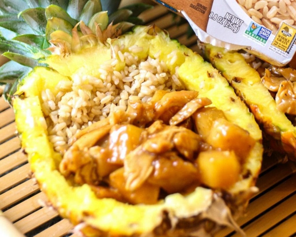 A pineapple half filled with rice and a savory chicken breast topping, presented on a bamboo mat.