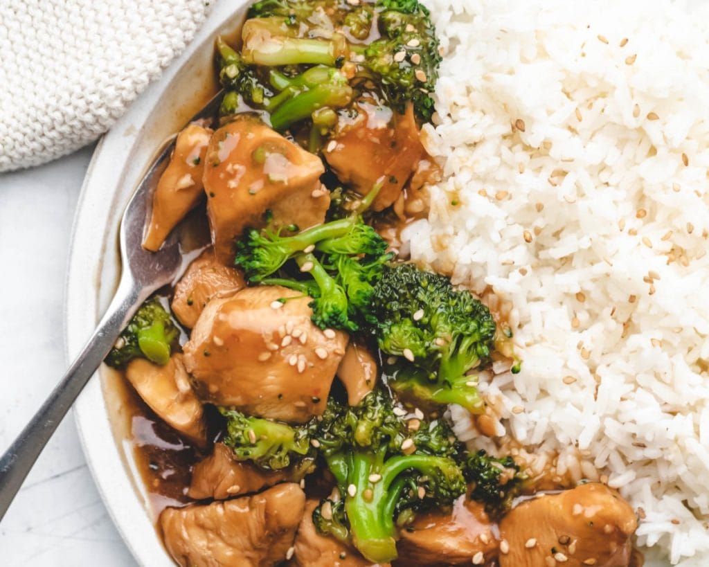 A plate of chicken breast and broccoli stir-fry served next to white rice, garnished with sesame seeds.
