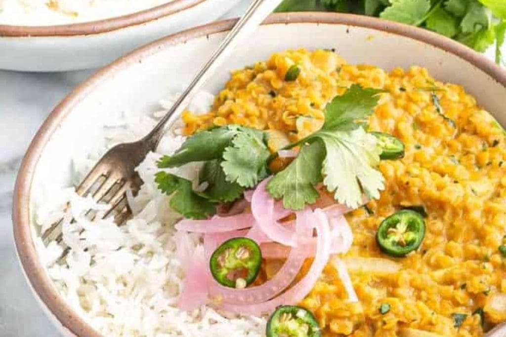 A bowl of lentil curry recipes served over rice, garnished with cilantro leaves, pickled onions, and sliced jalapeos.