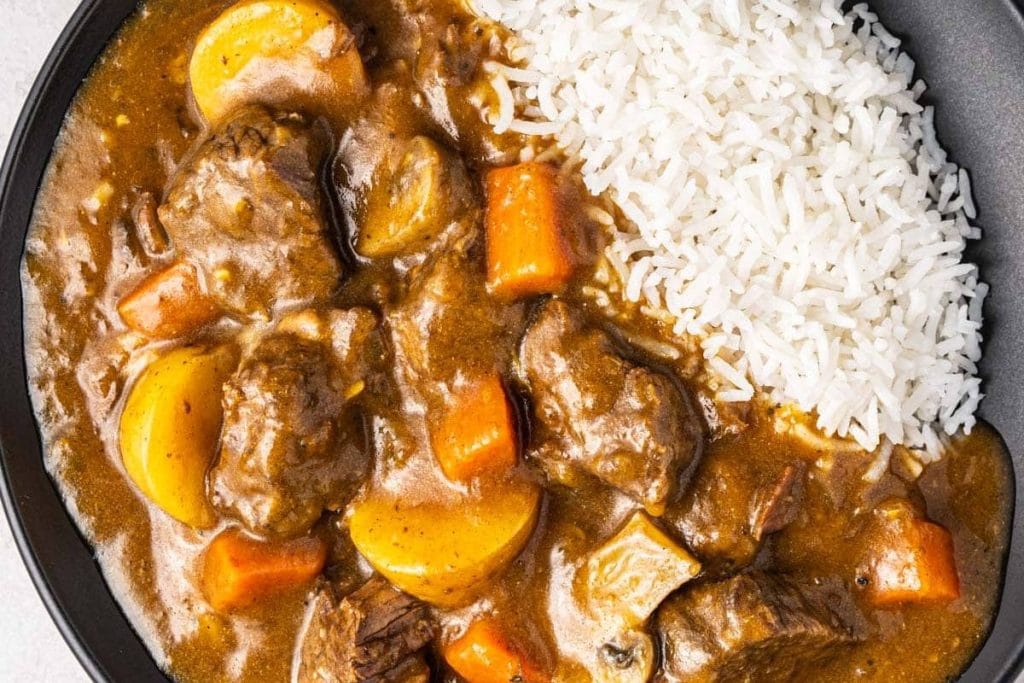Beef curry recipes with carrots and potatoes served alongside white rice in a black bowl.