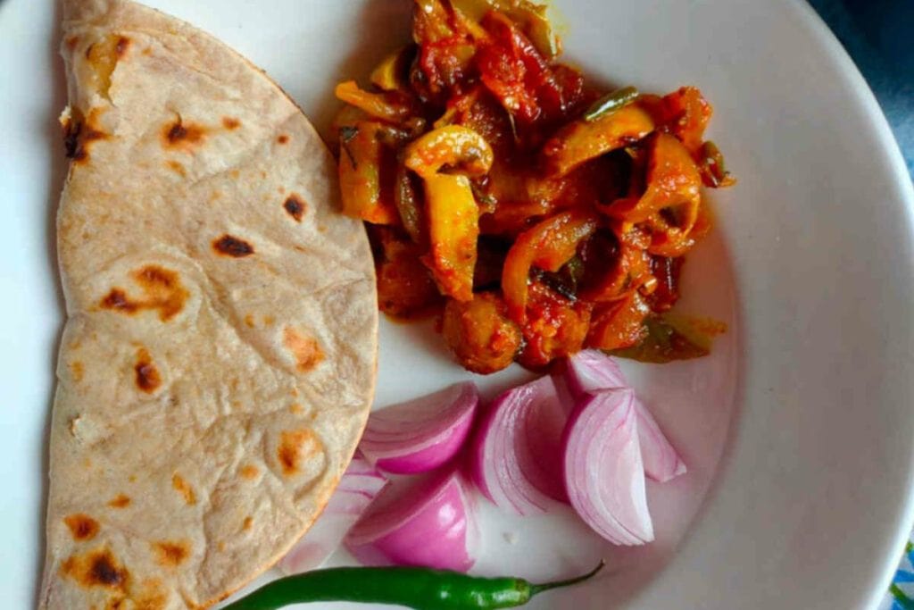 A plate of Indian food consisting of chapati, mixed vegetable curry recipes, sliced onions, and a green chili pepper.