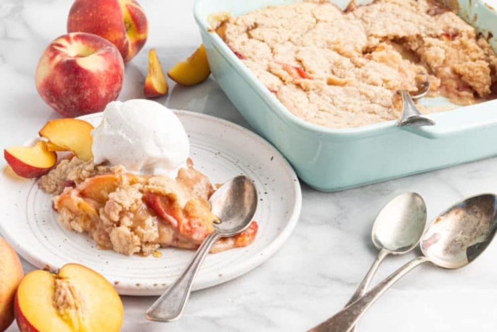 Spring desserts like peach cobbler served on a plate with a scoop of ice cream, alongside fresh peaches and a baking dish.