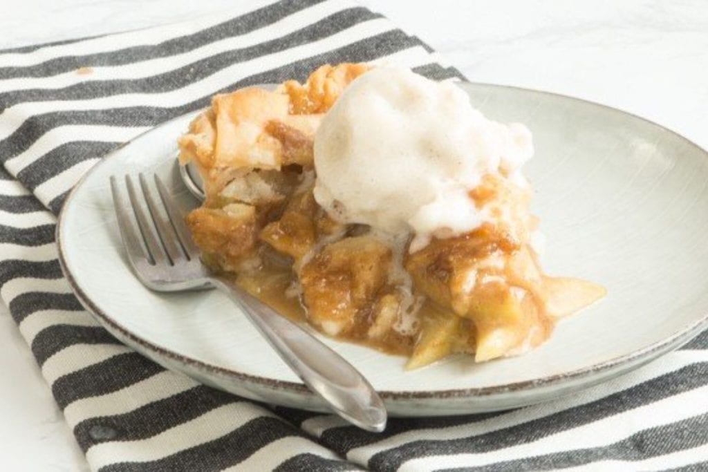 A serving of apple pie with a scoop of melting ice cream on a plate with a fork, on a striped napkin, perfect for spring desserts.