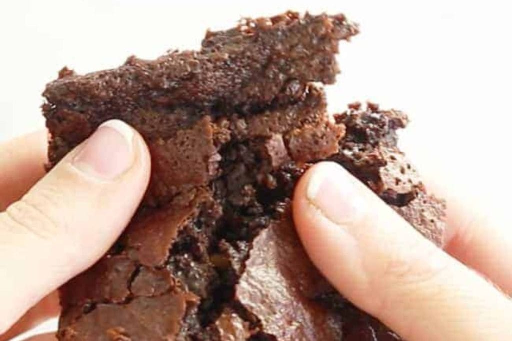 A close-up of a person's fingers holding a broken piece of a moist, dense chocolate brownie, perfect for spring desserts.