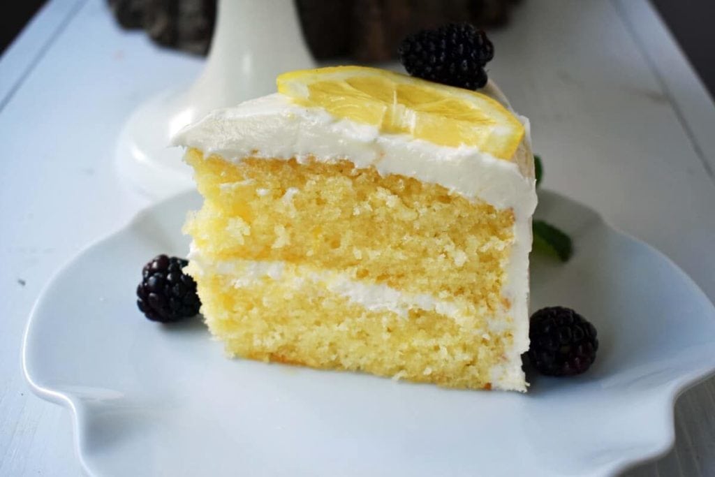 A slice of lemon cake with white frosting, perfect for spring desserts, garnished with a lemon slice and blackberries on a white plate.
