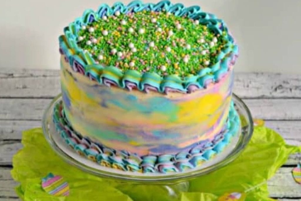 A colorful frosted spring cake with sprinkles on top displayed on a glass stand.