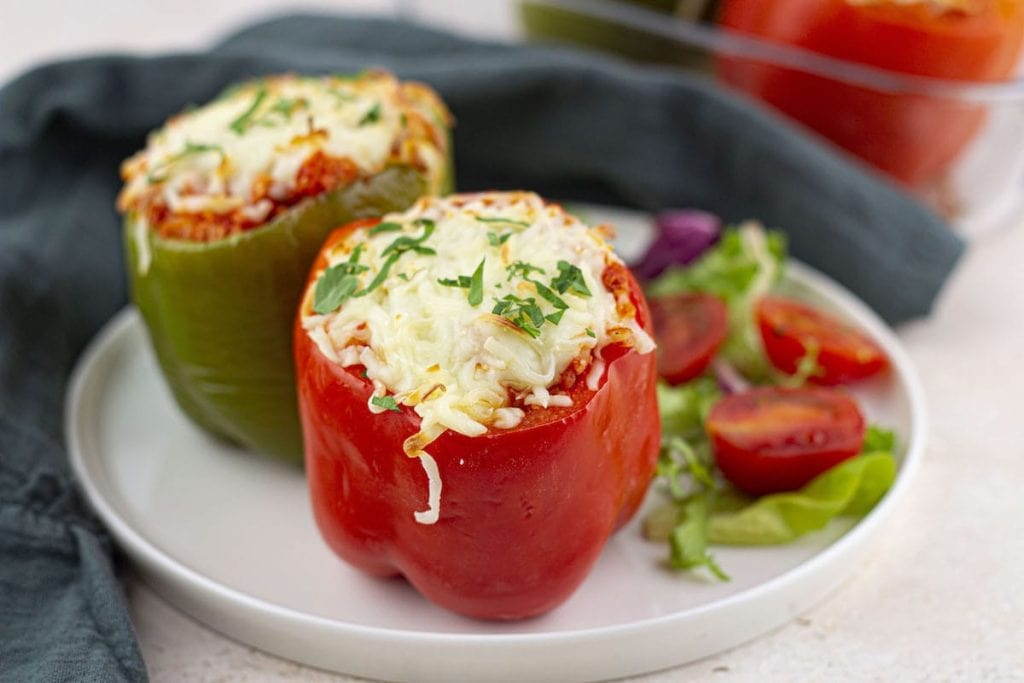Two stuffed bell peppers, one red and one green, topped with melted cheese and garnished with parsley, served on a white plate with salad on the side.