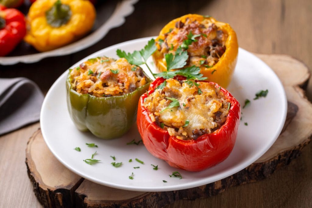 Three stuffed bell peppers (red, green, yellow) filled with rice and meat, garnished with parsley, on a wooden table.