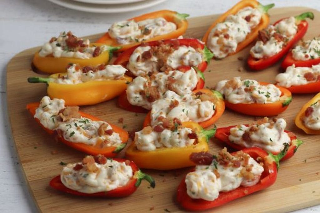 Stuffed bell peppers with cream cheese and topped with crispy bacon, garnished with herbs on a wooden cutting board.