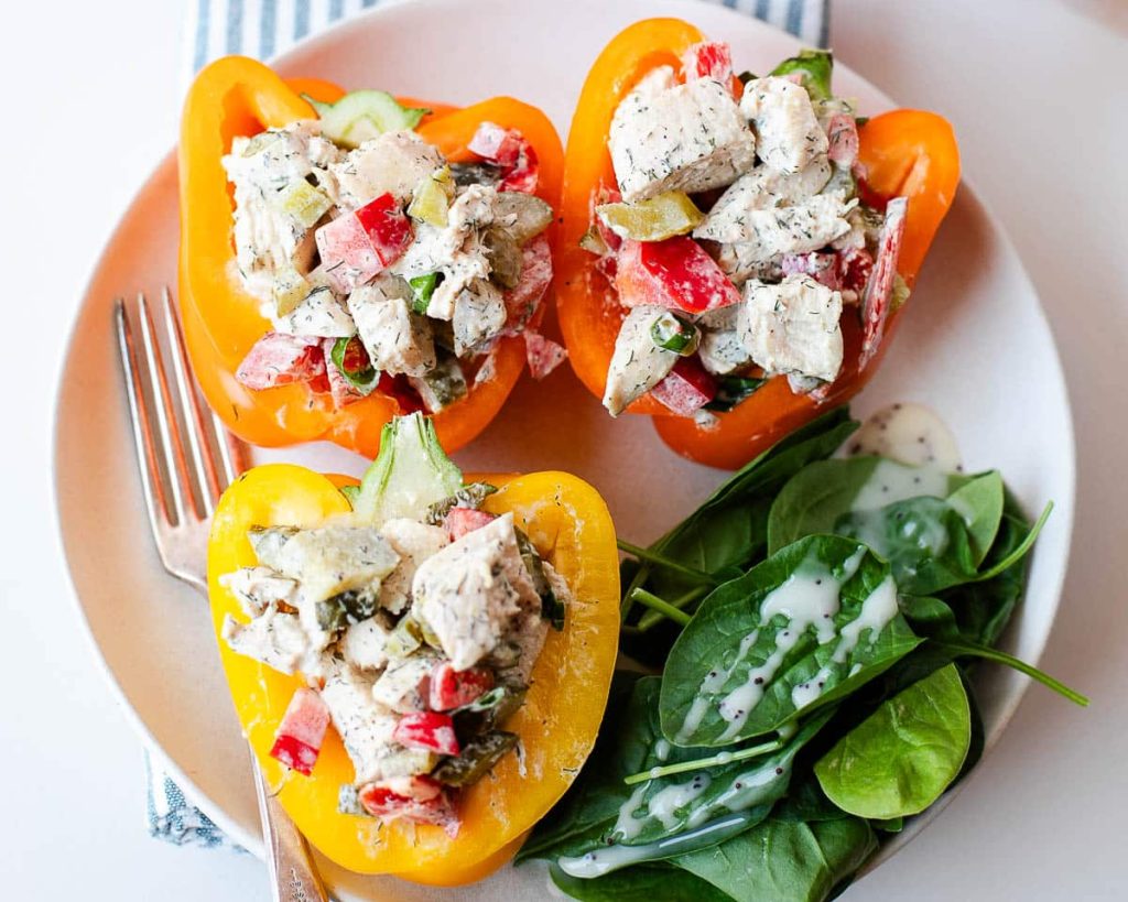 Two stuffed bell peppers with chicken salad, served on a white plate with a side of spinach, next to a striped napkin and a pepper package.