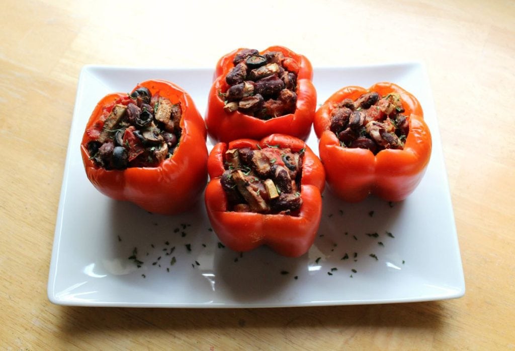 Four stuffed bell peppers on a white plate, filled with a mixture of diced vegetables and herbs, served on a wooden table.